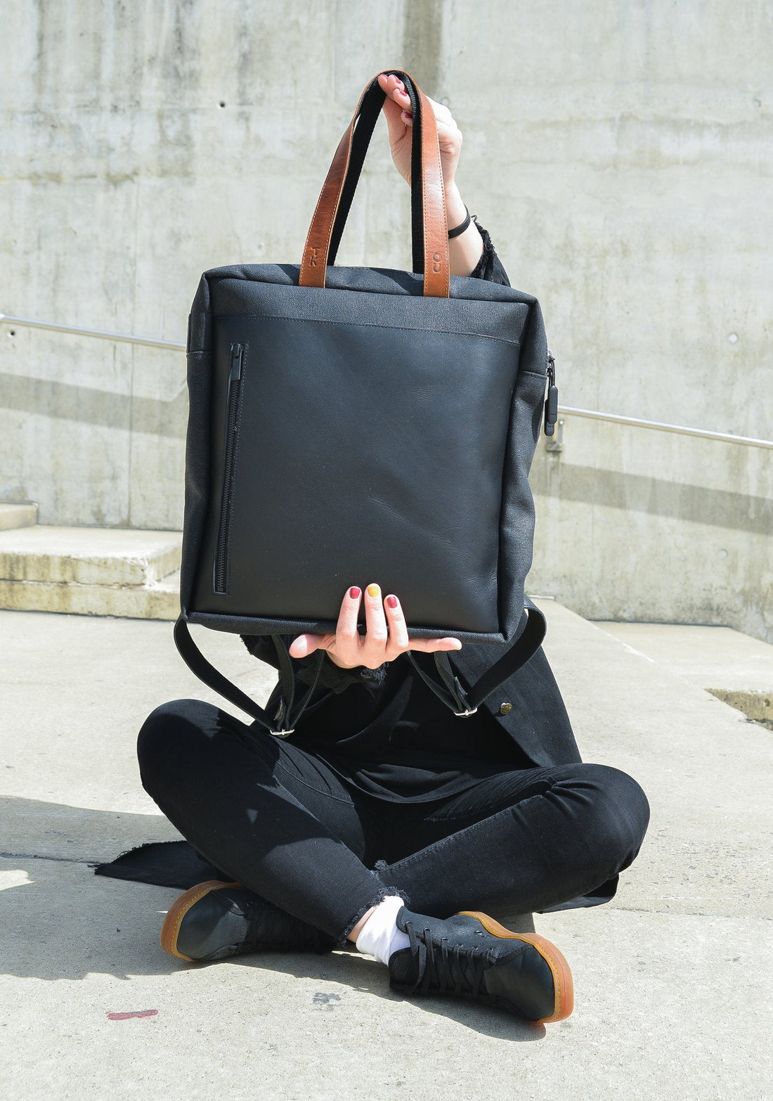 1. Unisex Stockholm Backpack in beige held by a model sitting outdoors, showcasing versatile design and leather details