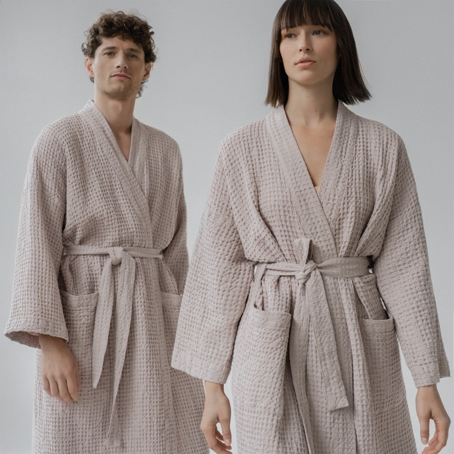 1. Man and woman wearing powder-colored honeycomb waffle bathrobes with adjustable waist ties and pockets, standing in a studio setting