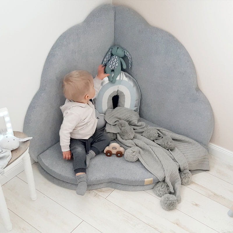 1. Toddler sitting on light grey cloud-shaped play mat in corner of room with toys and blanket