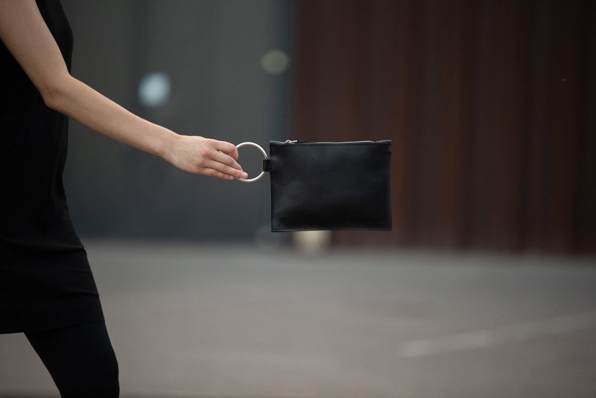 1. Woman holding PYKOK Clutch Nina with metal ring handle, showcasing sleek black leather design in urban setting