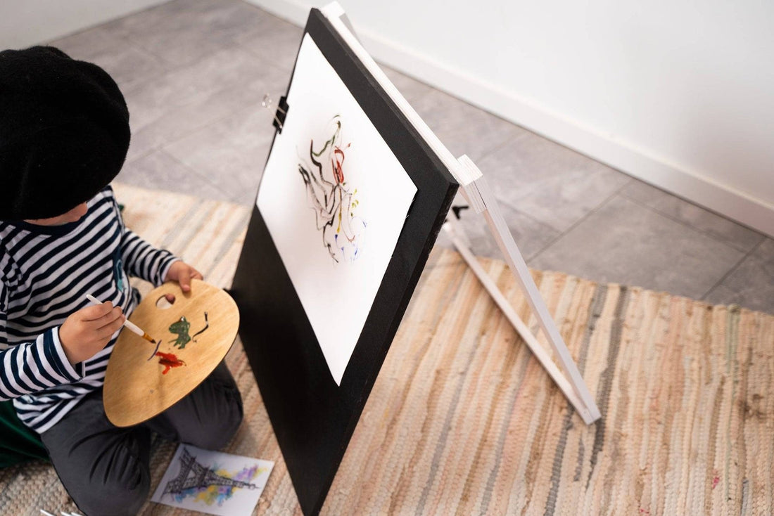 1. Child in striped shirt and beret painting on Ewart Woods black wooden chalkboard easel with art supplies on floor