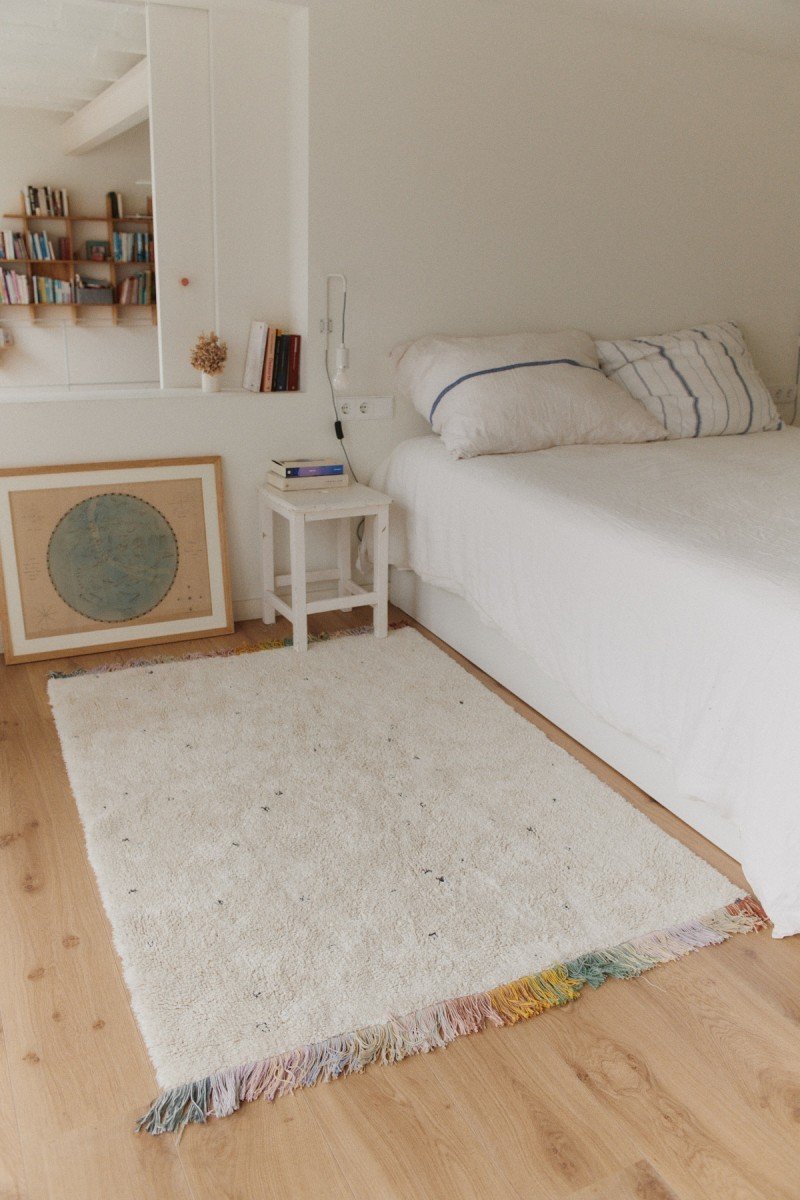 1. Lorena Canals Stardust washable rug in bedroom setting, placed beside a bed with white bedding, showcasing natural color and multicolored fringes
