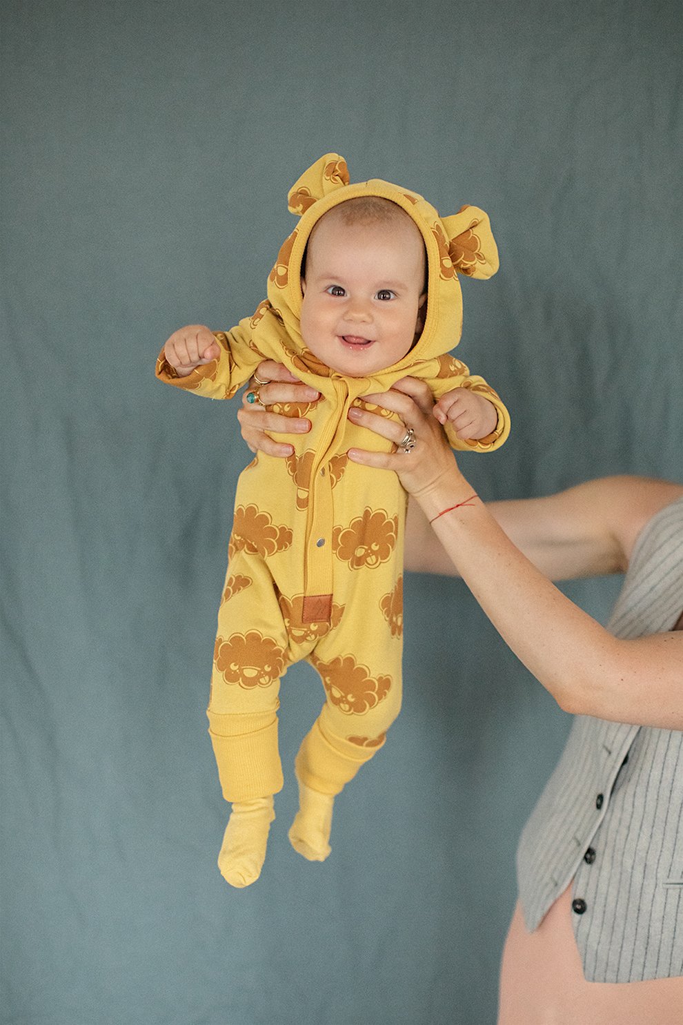 3. Baby held up wearing yellow jumpsuit with cloud print and hood with ears, smiling