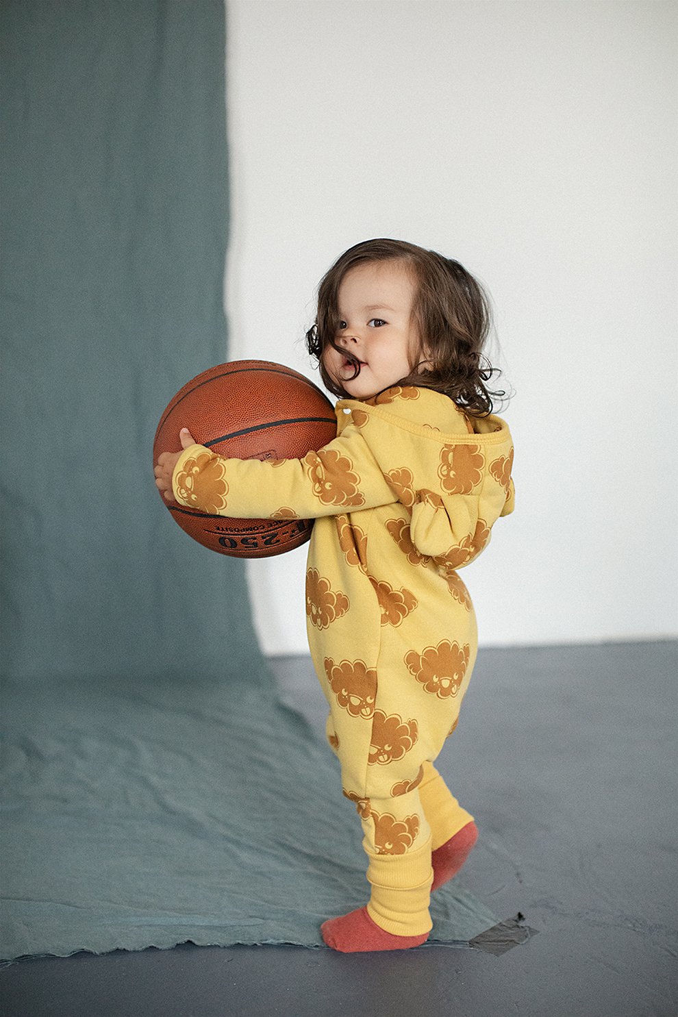 1. Toddler wearing yellow jumpsuit with cloud print and hood with ears, holding basketball indoors