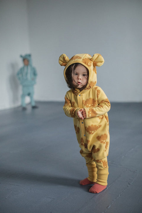 4. Child in yellow jumpsuit with cloud print and hood with ears, standing indoors with another child in background