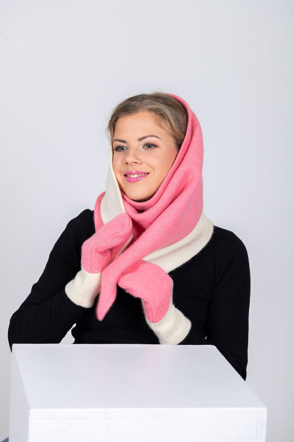 2. Woman draping HERTTA pink and white wool triangle shawl over shoulders with matching mittens, studio setting
