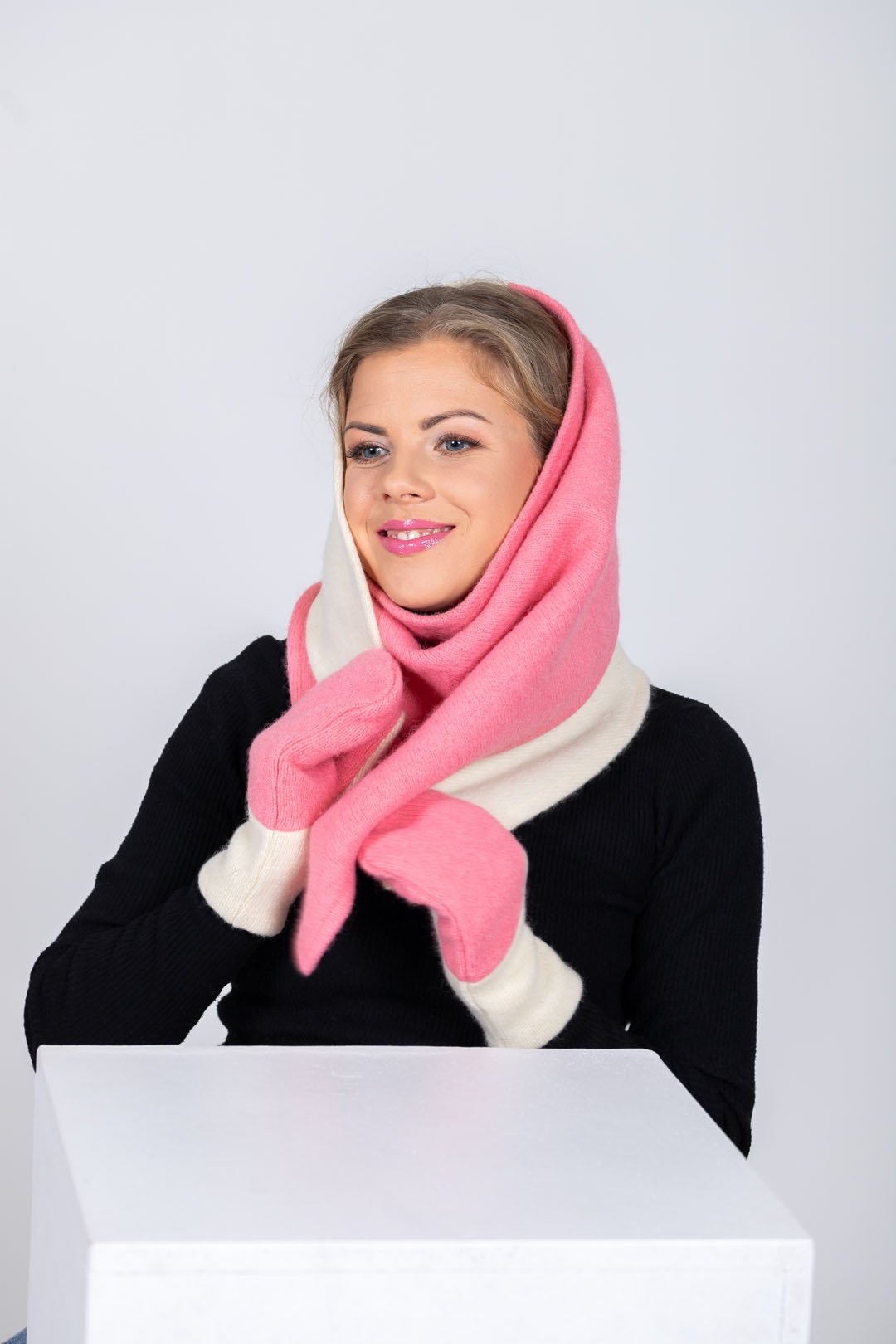 2. Woman draping HERTTA pink and white wool triangle shawl over shoulders with matching mittens, studio setting