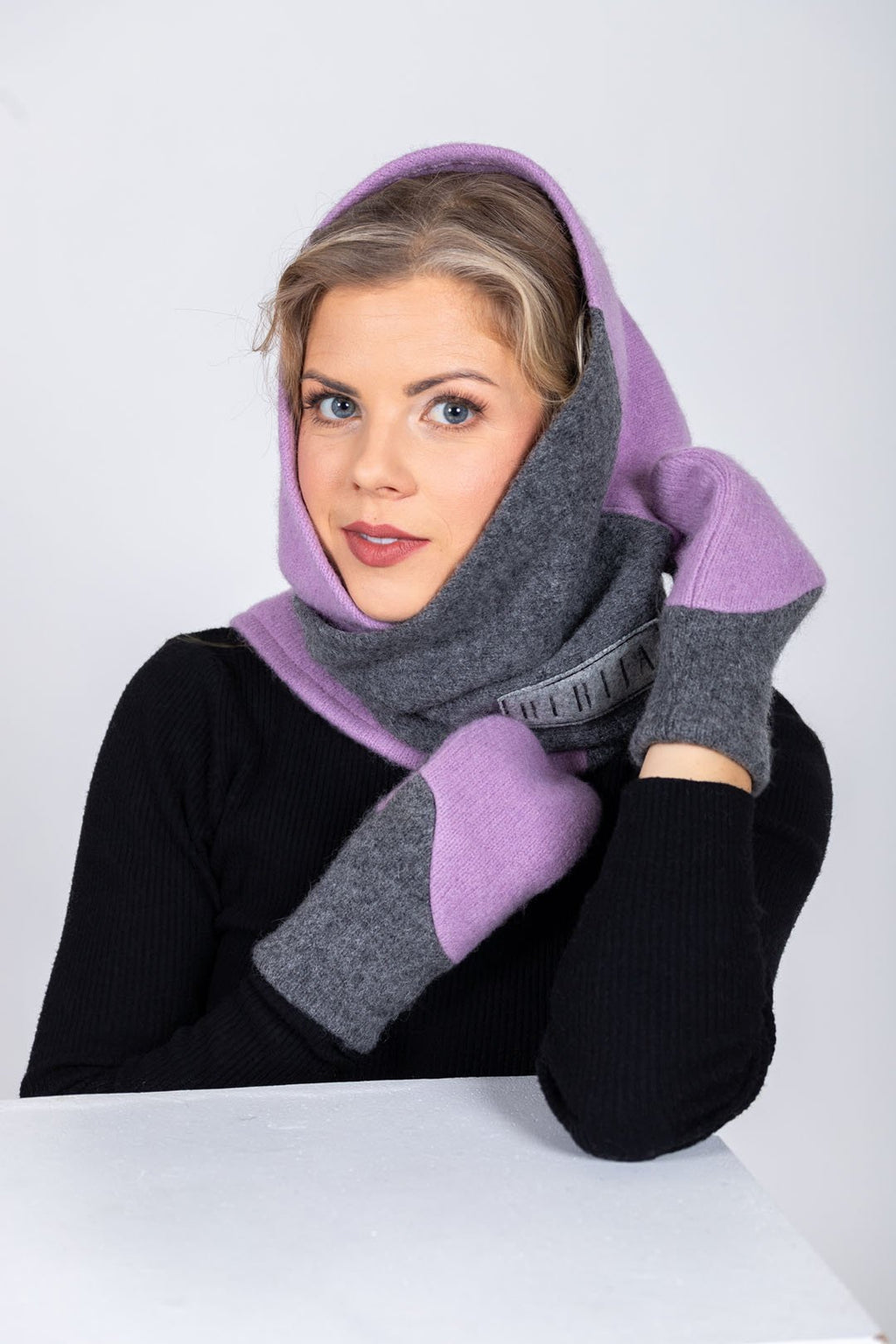 5. Woman wearing HERTTA lilac and medium grey wool triangle shawl as a hood with embroidered logo, in studio setting