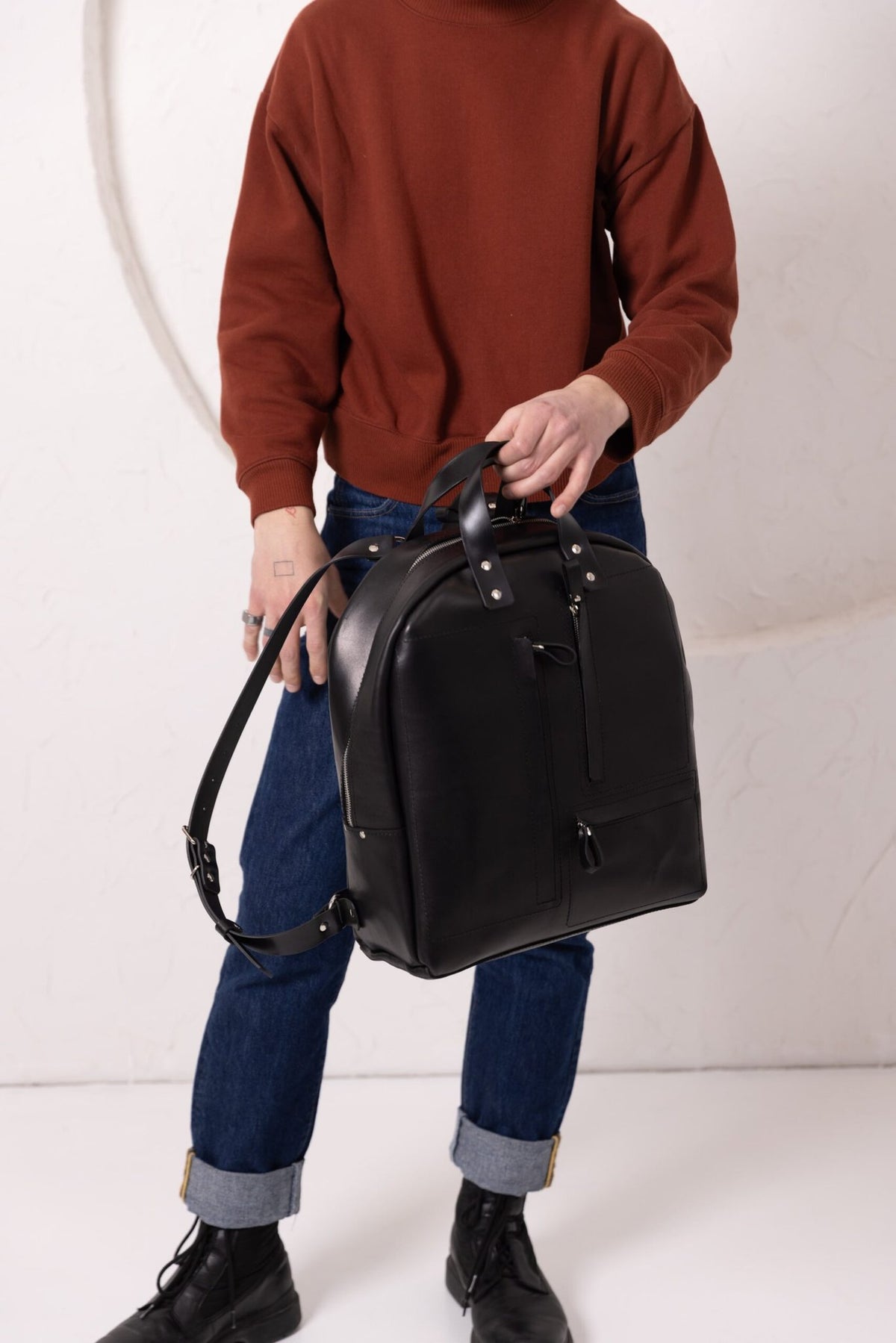 7. Man holding TOKS VISOKS GORDON black leather backpack, showcasing adjustable straps in studio setting