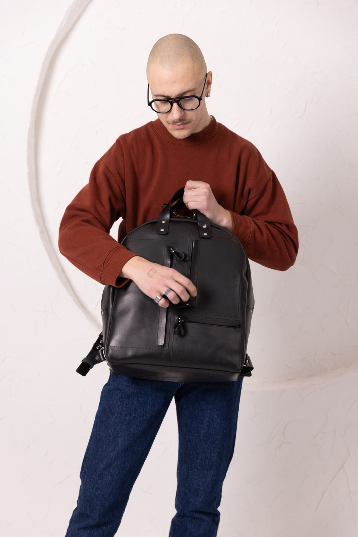 13. Man holding TOKS VISOKS GORDON black leather backpack, emphasizing front zipped pockets in studio setting