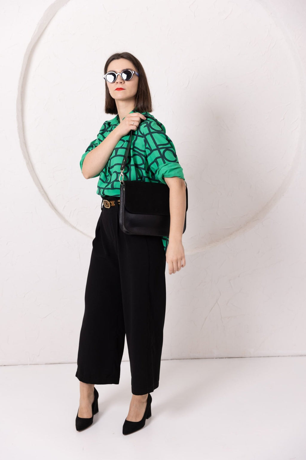 3. Woman showcasing TOKS VISOKS ASTORIA black suede handbag with adjustable strap, styled with green top and black pants