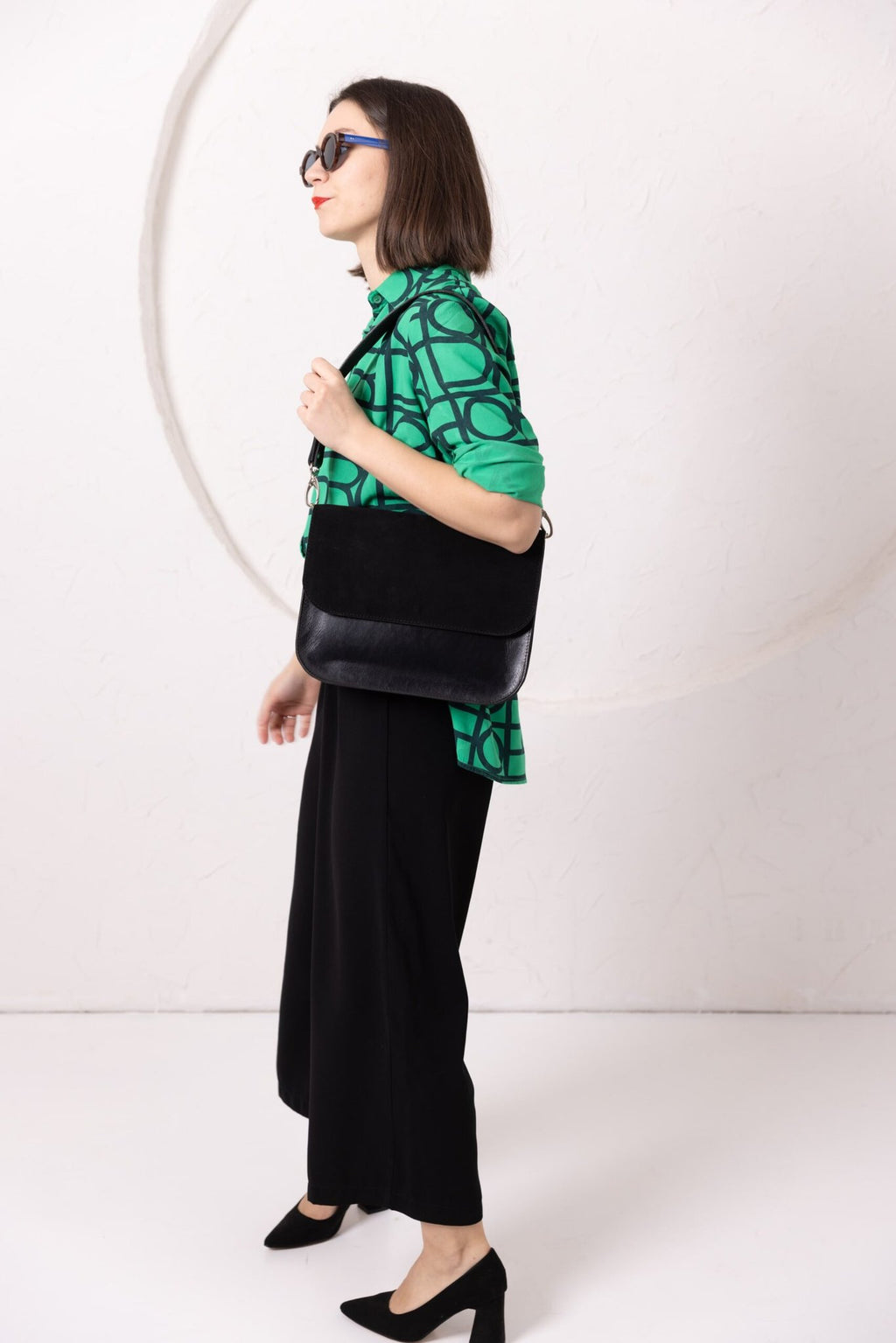 4. Side view of woman wearing TOKS VISOKS ASTORIA black suede handbag, styled with green top and black pants