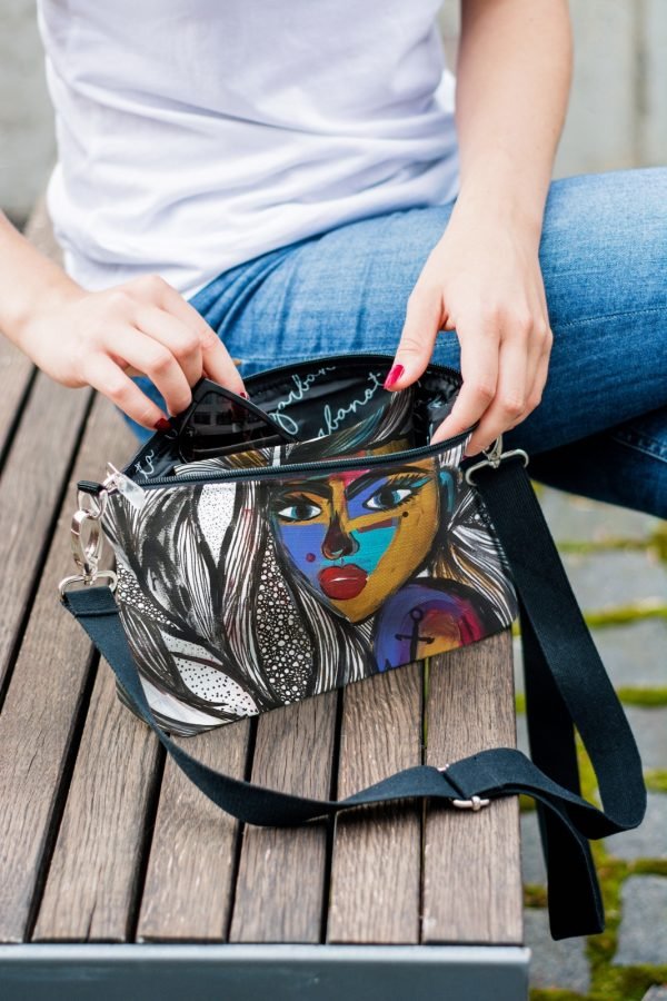 1. Women's shoulder bag with mermaid artwork by Dovilė Aleksonė, shown open with hand accessing interior on bench