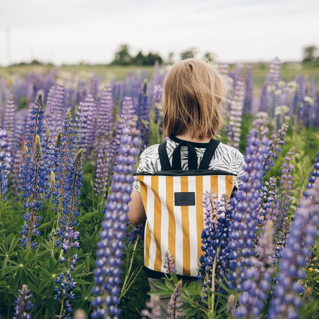 1. Child wearing Muni striped mustard and white backpack in a field of flowers