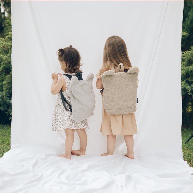 3. Back view of two young girls wearing Muni beige teddy backpacks, standing on white fabric