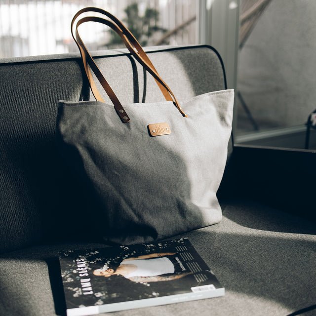 5. Grey denim tote bag by Muni with tan leather handles, placed on grey sofa with magazine in lifestyle setting