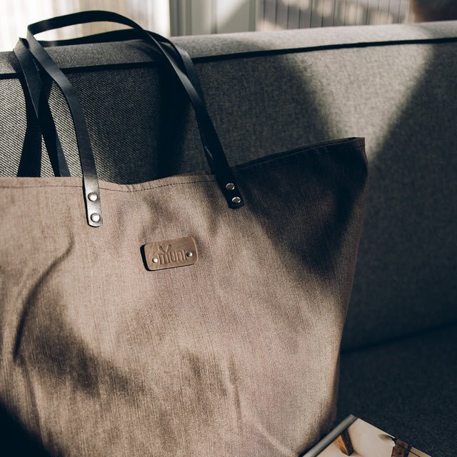 8. Muni brown denim tote bag with black leather handles resting on a grey sofa, showing brand logo