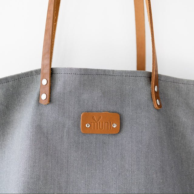 7. Muni grey denim tote bag with tan leather handles, hanging on wooden hook in studio setting
