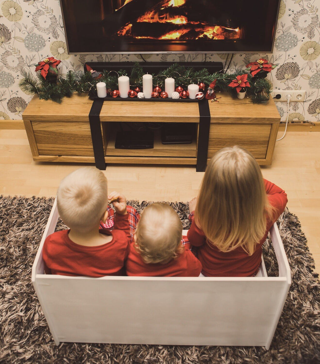 2. Three children in red clothing sitting in a white box watching a TV with a festive setup