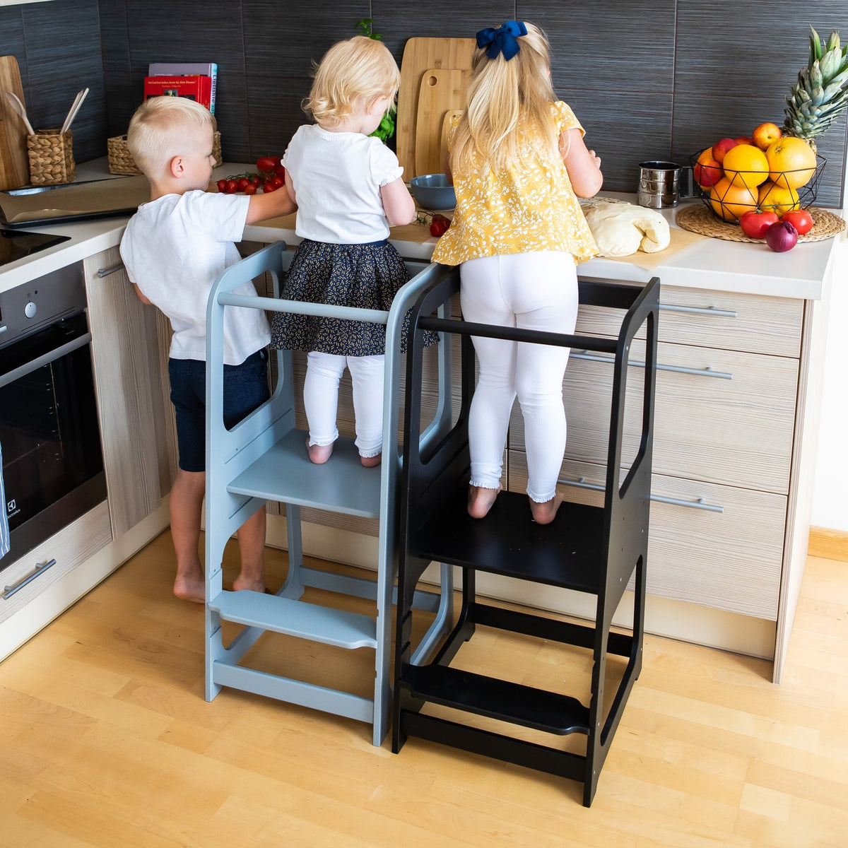 21. Three children using blue and black Montessori helper tower step stools in kitchen