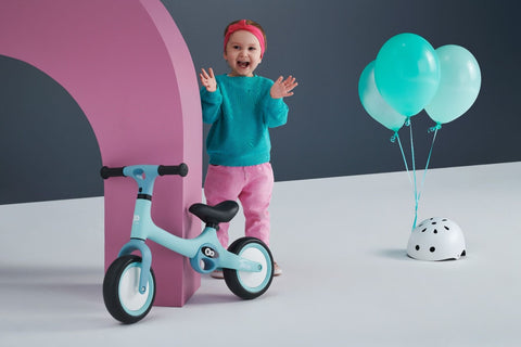 1. Child standing next to mint green Kinderkraft TOVE balance bike with balloons and helmet in playful setting