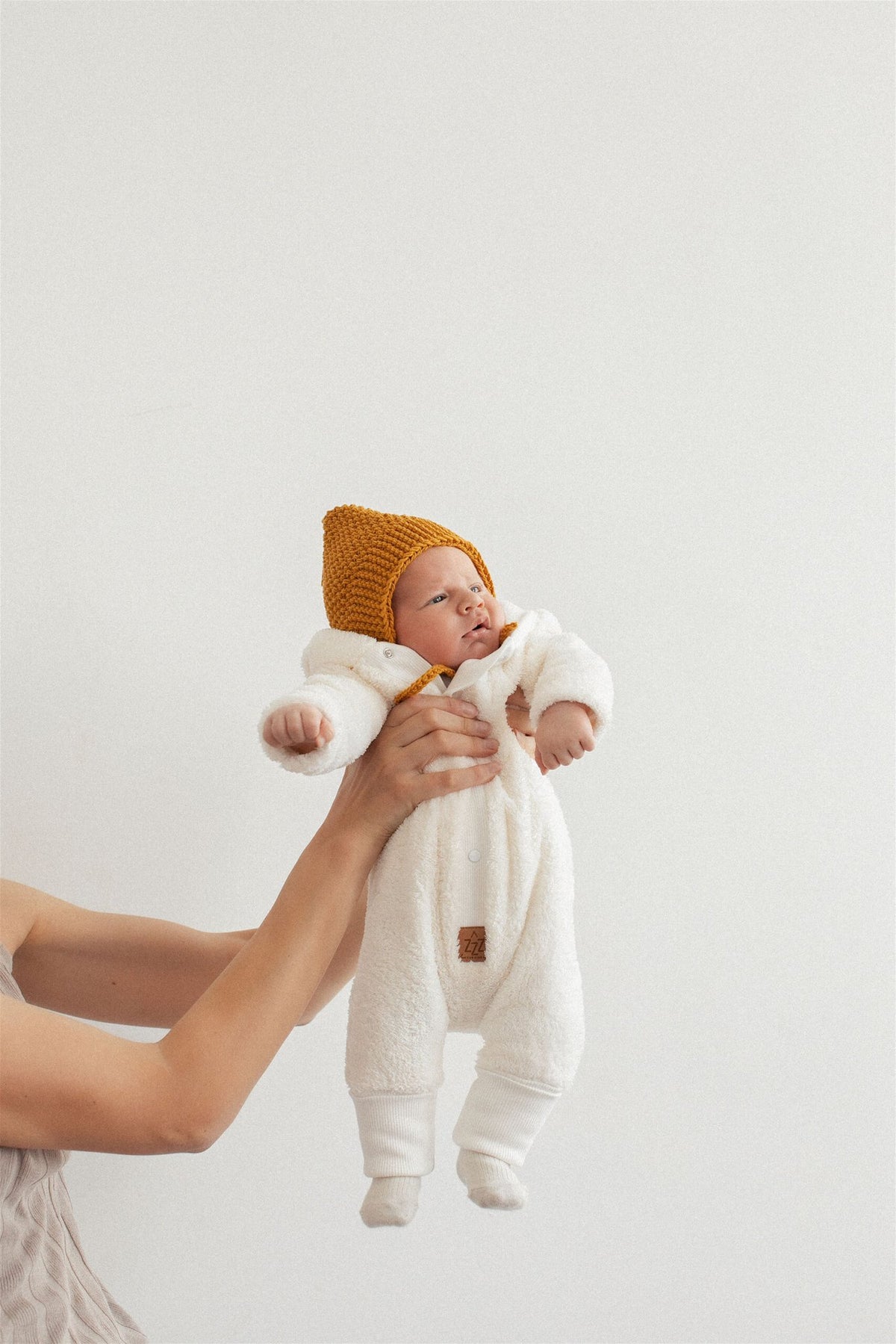 2. Baby held up wearing fuzzy ecru jumpsuit with bear ears hood and mustard knit hat