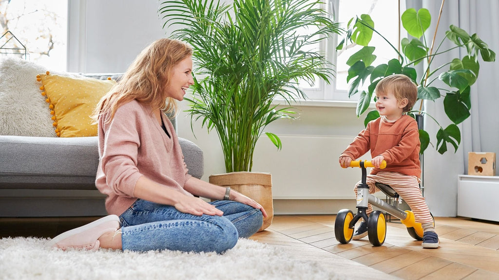 8. Child riding yellow Kinderkraft CUTIE balance bike with parent watching in living room