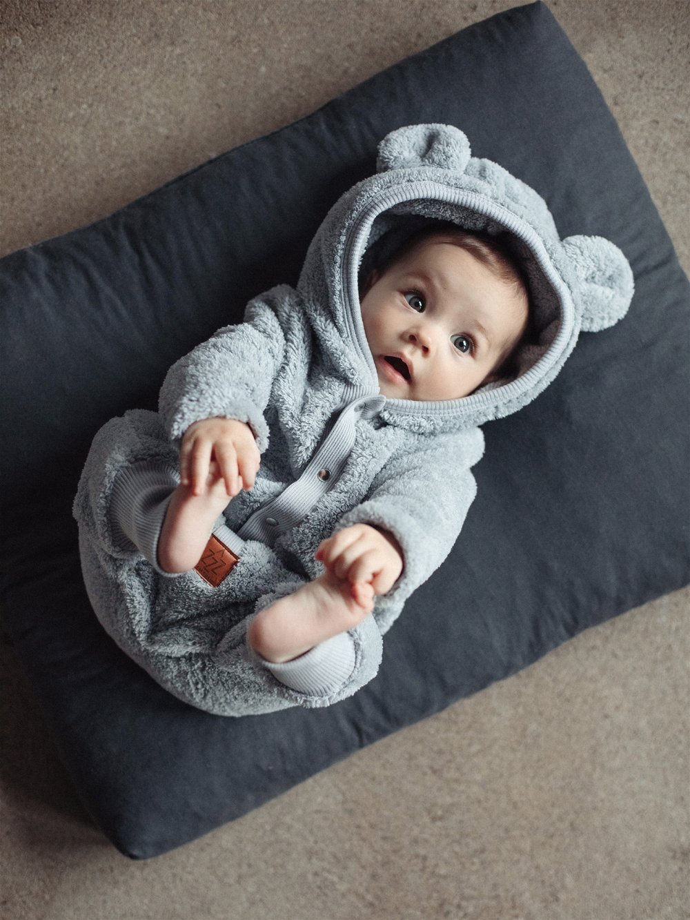 1. Baby wearing fuzzy grey eared jumpsuit lying on dark cushion, showcasing playful hood with ears