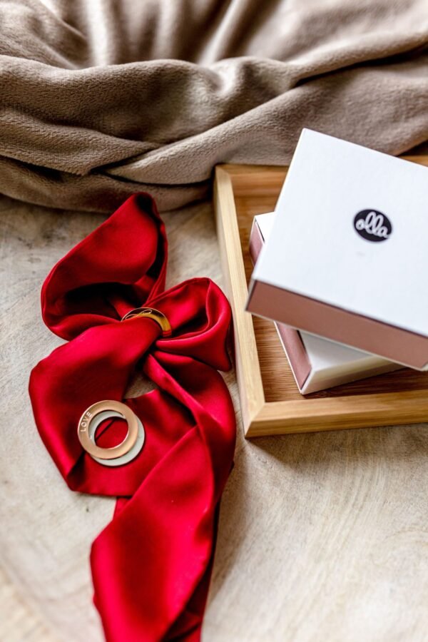 3. Flat lay of Olla Silk Ribbon Bracelet in dark red with gold-plated ring, displayed with branded packaging