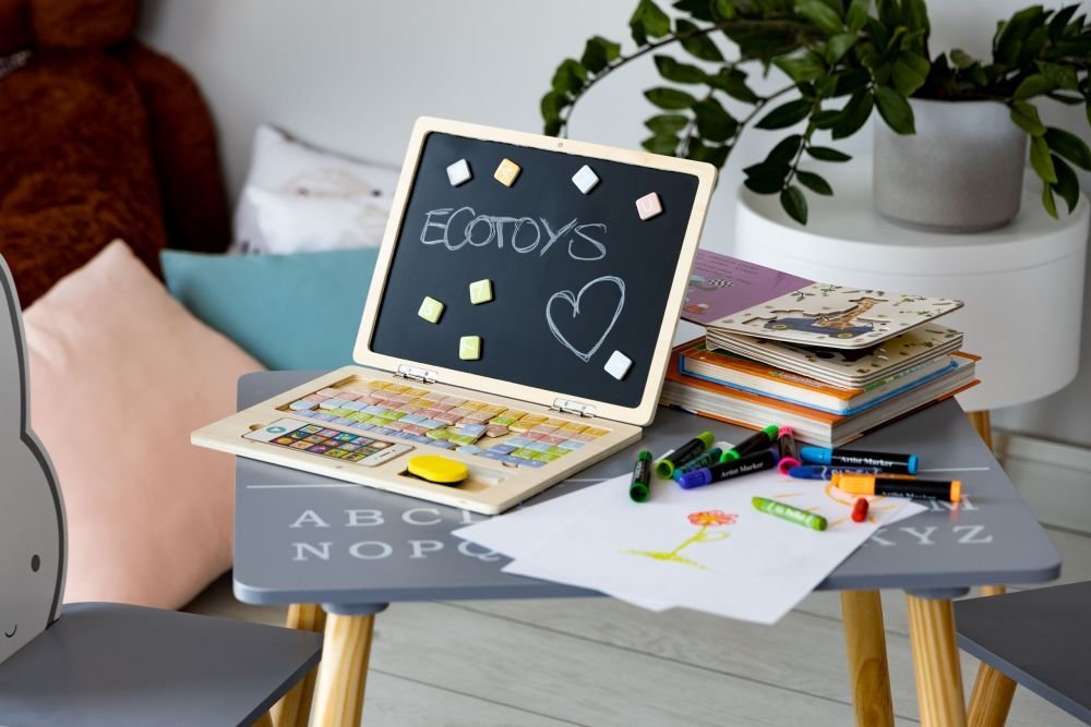 1. Ecotoys wooden laptop and phone set on a children's table with magnetic letters and chalk drawings, surrounded by colorful books and crayons