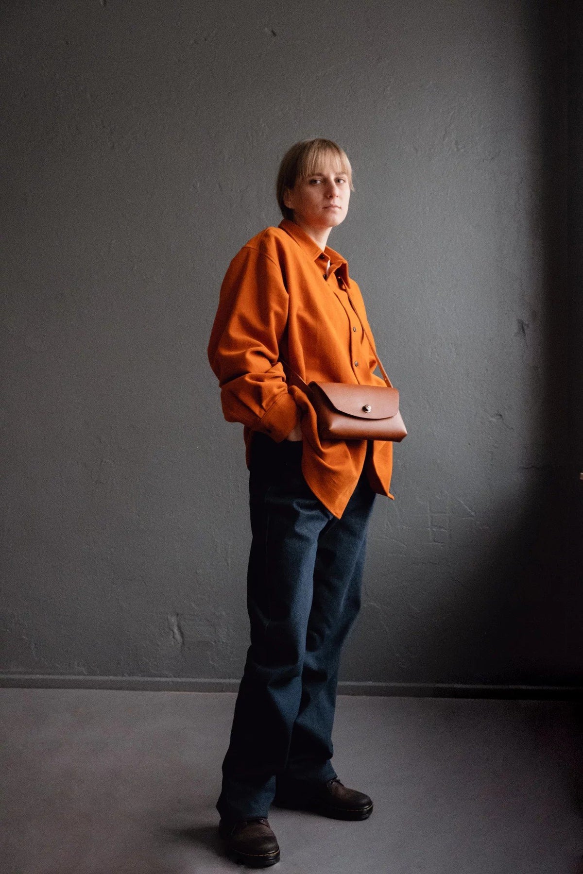 3. Full view of woman wearing rust brown leather clutch by Mirgoods, paired with orange shirt and jeans
