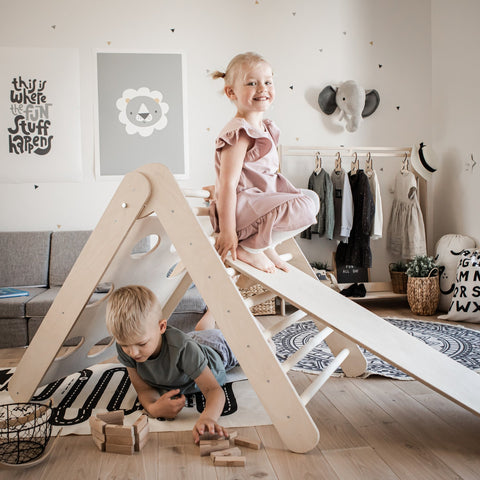 1. Two children playing on Montessori climbing triangle with ramp in a cozy playroom, girl sliding down ramp, boy playing with blocks underneath.