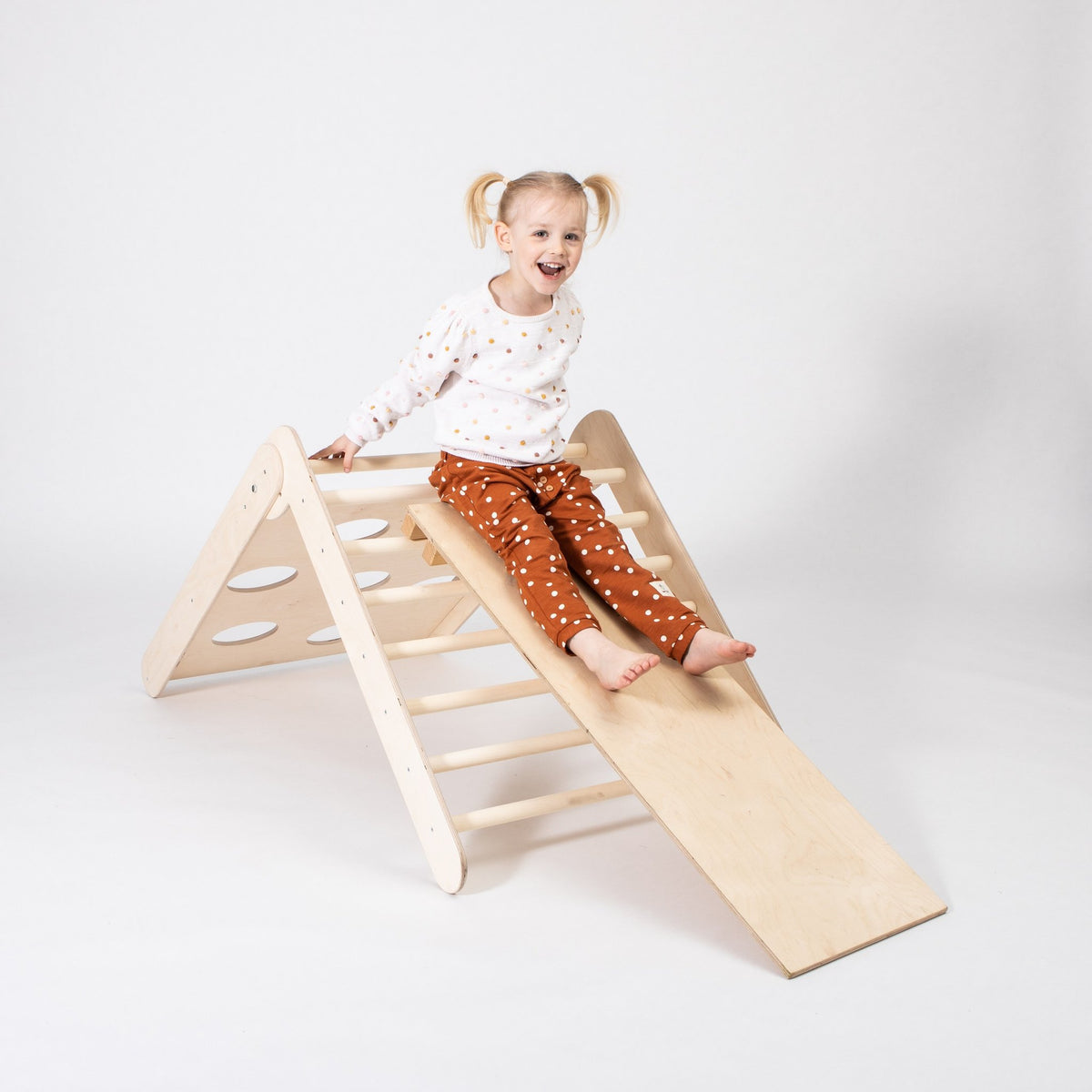 1. Girl sliding down Montessori climbing triangle with ramp in studio setting, wearing polka dot outfit, smiling.