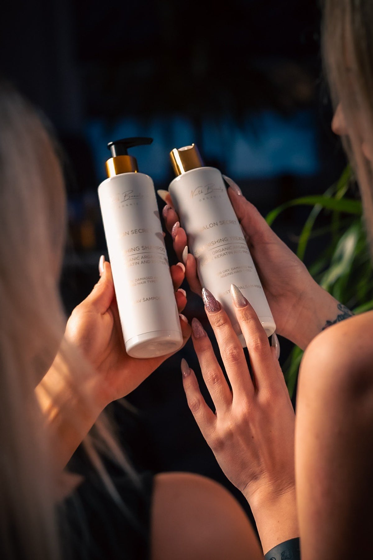 1. Two women holding North Beauty shampoo and treatment bottles in a dimly lit room, showcasing product labels and gold caps