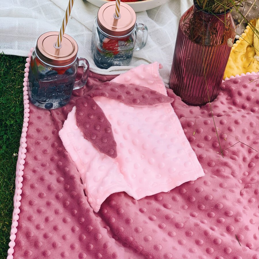 3. Close-up of dusty rose 2in1 blanket with bunny ears comforter on grass, next to drinks and vase