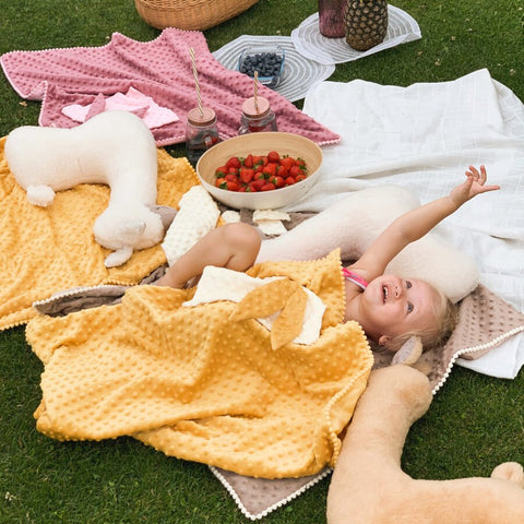 1. Child lying on grass covered with mustard 2in1 blanket with baby comforter, surrounded by plush toys and picnic items