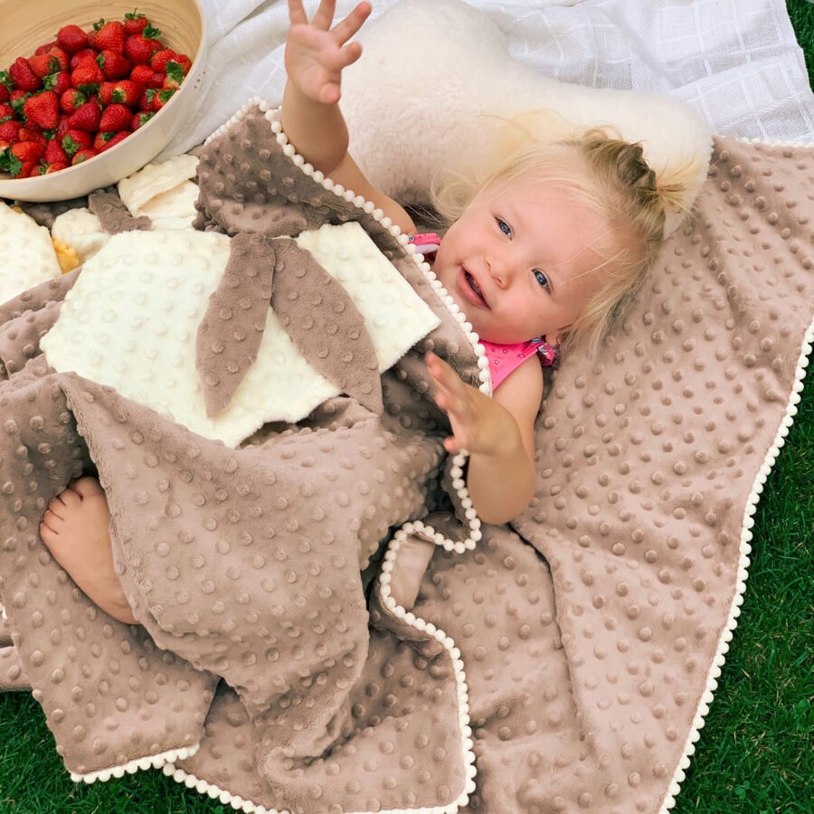 1. Baby lying on a beige textured blanket with bunny ears comforter, surrounded by strawberries and pillows on grass