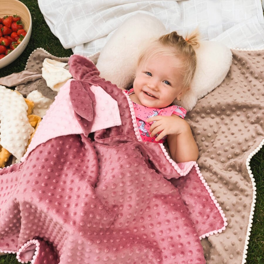 1. Smiling toddler wrapped in dusty rose 2in1 blanket with bunny ears comforter, lying on grass with picnic setup