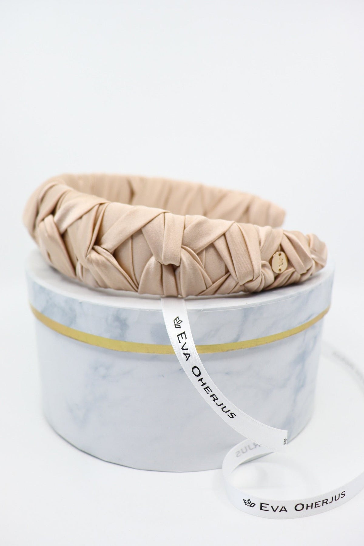 3. Side view of Eva Oherjus Jenna Mulberry Silk Knotted Headband in light beige with metal logo tag, on luxury hat box