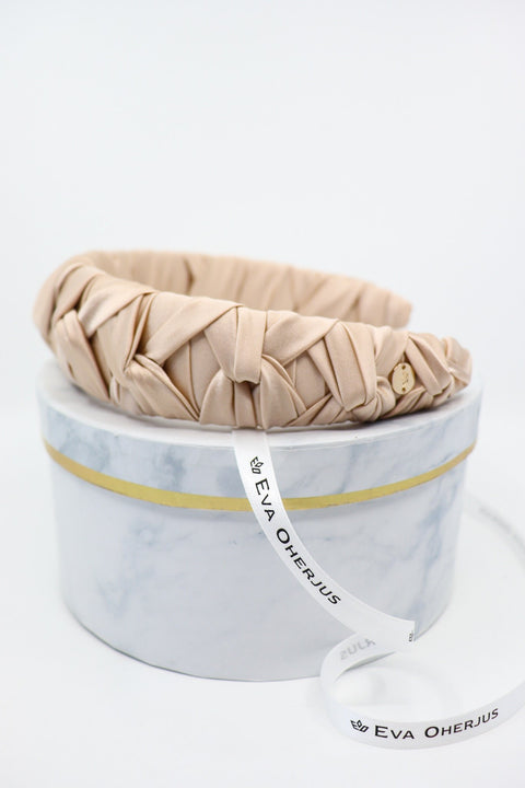 3. Side view of Eva Oherjus Jenna Mulberry Silk Knotted Headband in light beige with metal logo tag, on luxury hat box