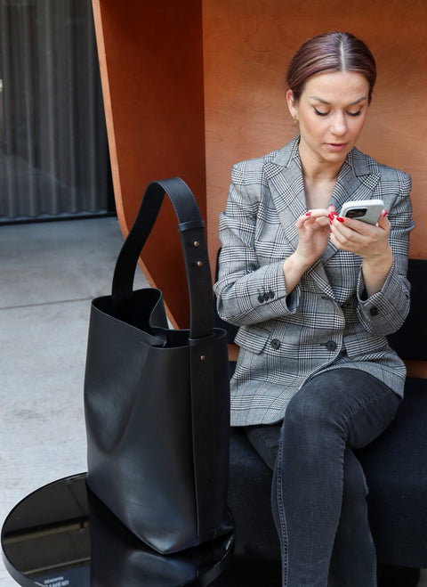3. Woman sitting with Tairi Roosve black leather tote bag, using phone, highlighting bag's versatility and style