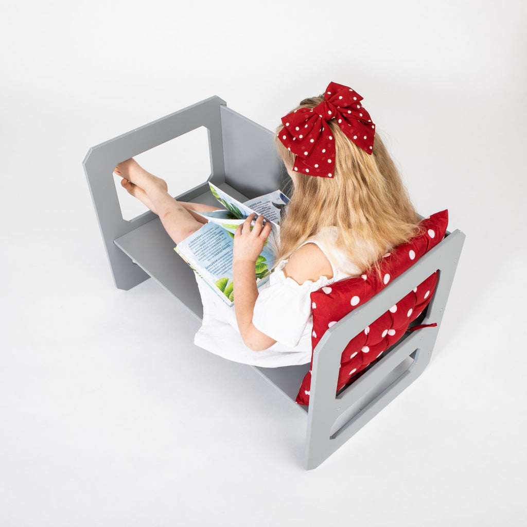 9. Girl with red bow reading on a grey Montessori chair with a red cushion on a white background