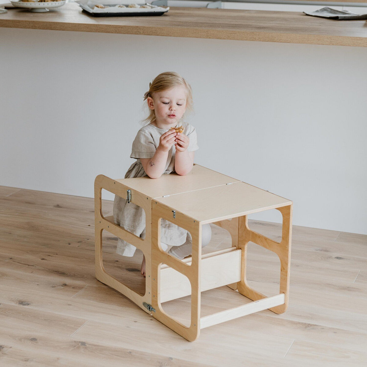 8. Child sitting at natural wood kitchen tower in table mode, enjoying a snack in a modern kitchen