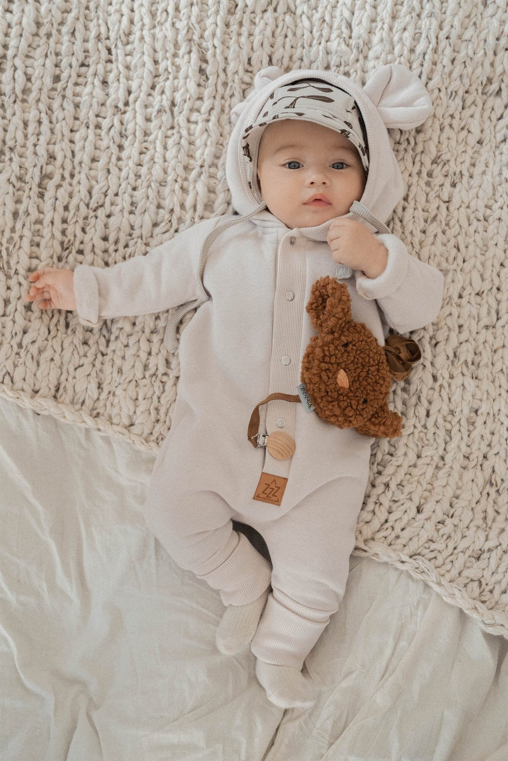 1. Baby wearing sand-colored eared jumpsuit with hood, lying on a knitted blanket, holding a plush toy