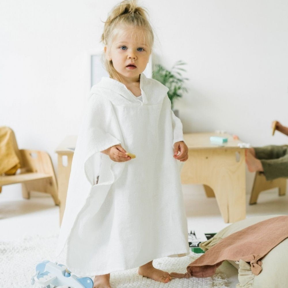 1. Child wearing white linen waffle poncho standing in a bright playroom