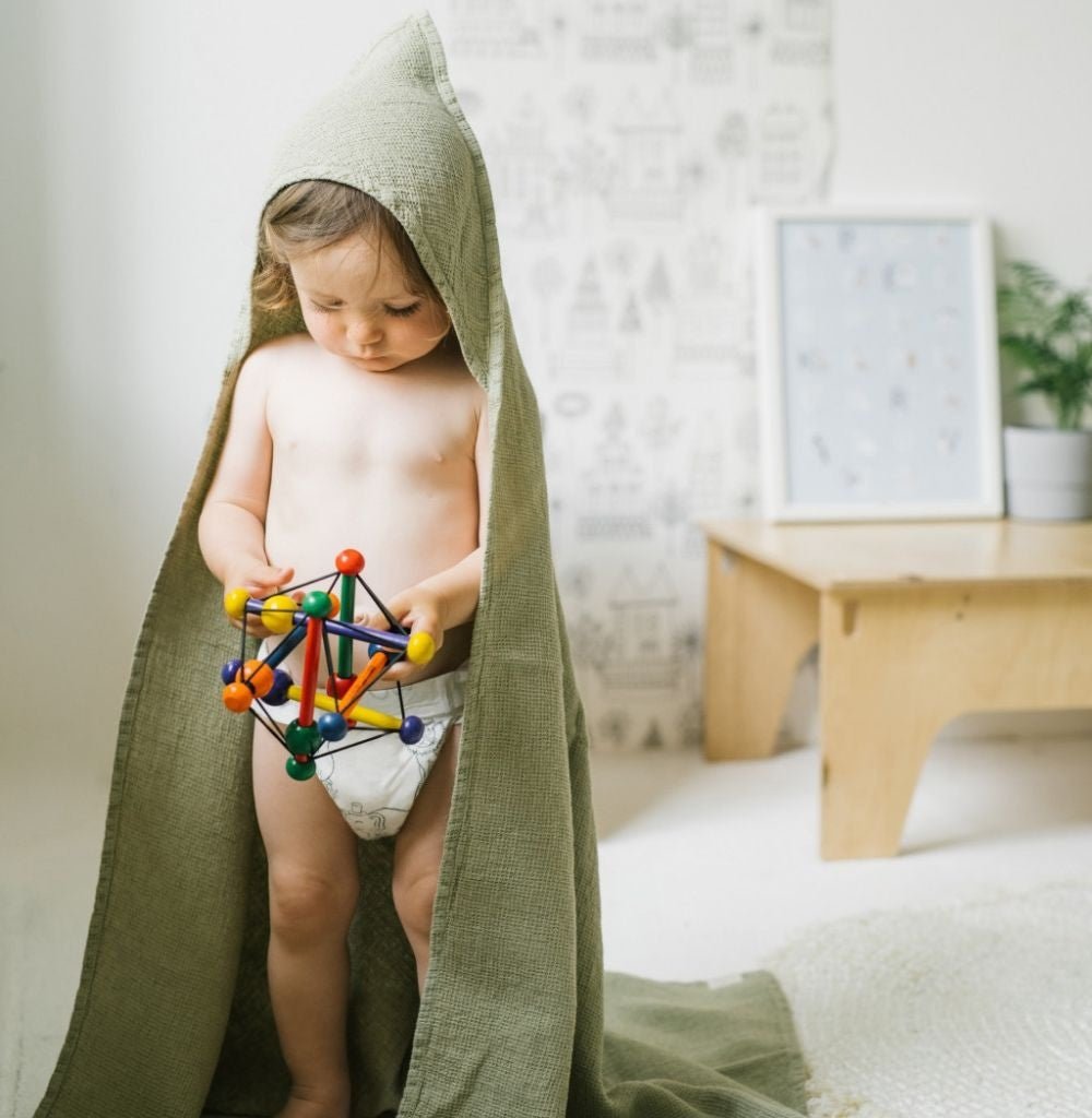 1. Child wearing sage green linen hooded towel playing with colorful toy in a cozy room