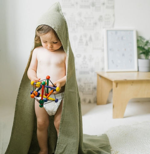 1. Child wearing sage green linen hooded towel playing with colorful toy in a cozy room