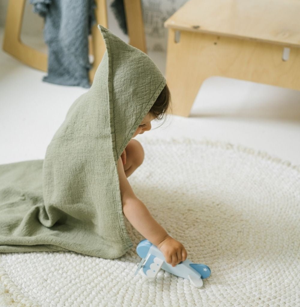 1. Child wearing a green linen hooded towel playing with a toy airplane on a round rug