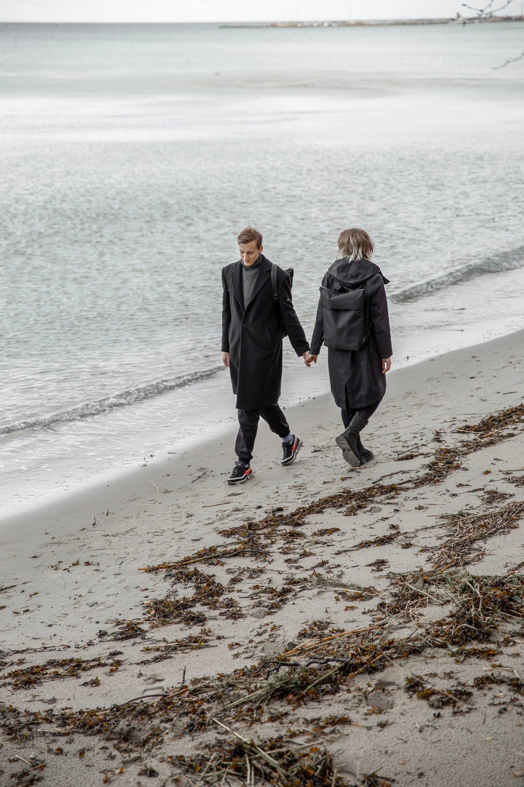 3. Unisex Nordhale HØLAR Weatherproof Backpack worn by male and female models holding hands on a beach, emphasizing style and functionality