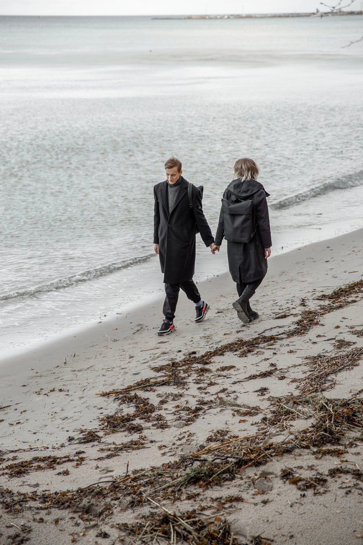 3. Unisex Nordhale HØLAR Weatherproof Backpack worn by male and female models holding hands on a beach, emphasizing style and functionality